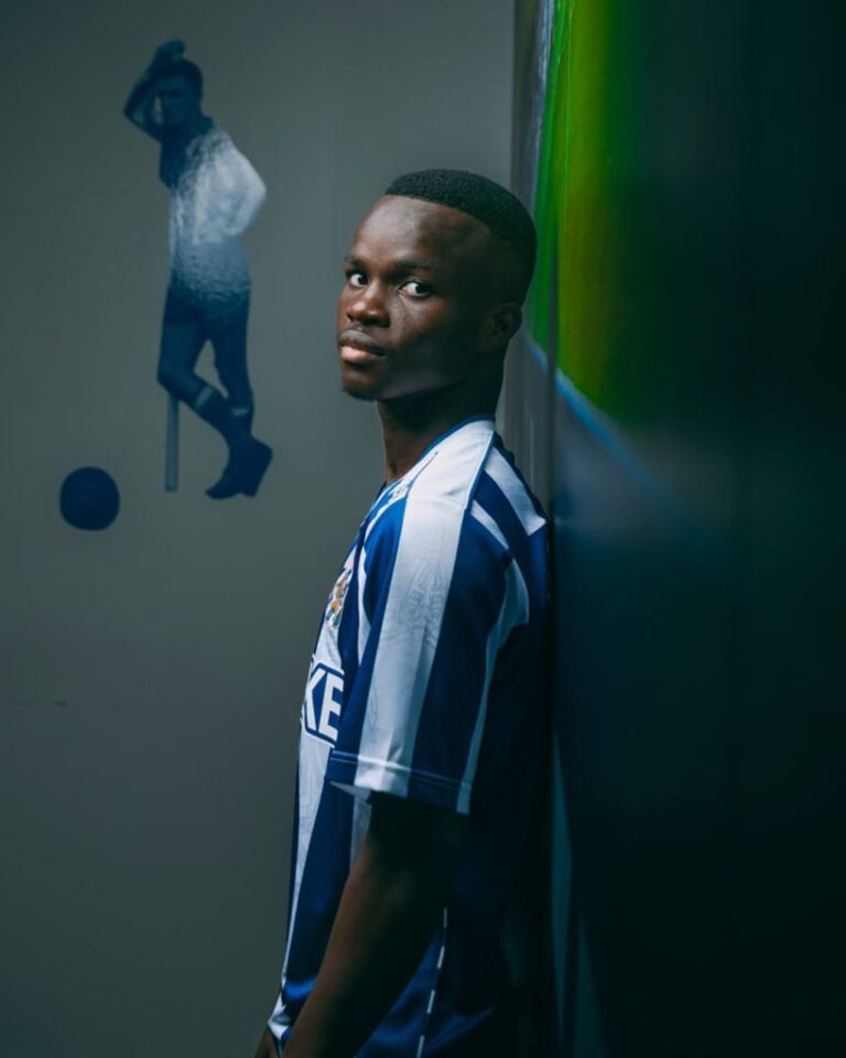 Young man in a blue-and-white striped soccer jersey leans against a wall, glancing at the camera in a dim hallway.