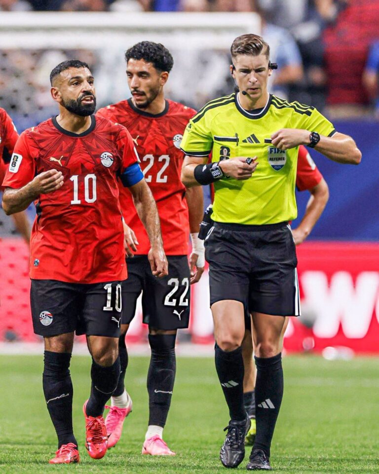 Soccer players in red jerseys walking on the field beside a referee in a bright yellow shirt.