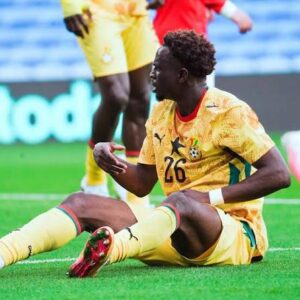 Soccer player in a yellow kit with number 26 sitting on the grass during a match, with arms gesturing and other players blurred in the background.