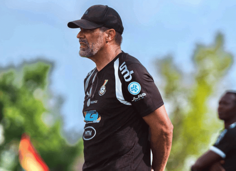 Man wearing a black cap and black team shirt with logos stands outdoors, looking to the left, with blurred trees in the background.