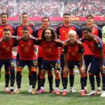 Spain national soccer team players pose for a group photo on the field in red and blue kits, with a green-clad goalkeeper on the right and a packed stadium in the background.