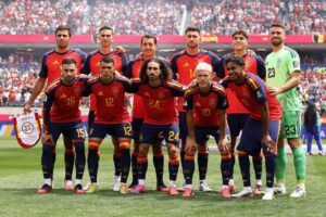 Spain national soccer team players pose for a group photo on the field in red and blue kits, with a green-clad goalkeeper on the right and a packed stadium in the background.