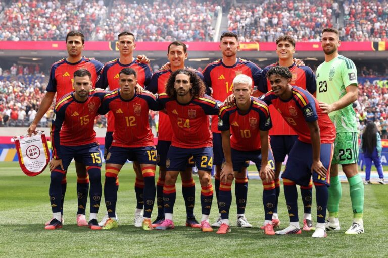 Spain national soccer team players pose for a group photo on the field in red and blue kits, with a green-clad goalkeeper on the right and a packed stadium in the background.
