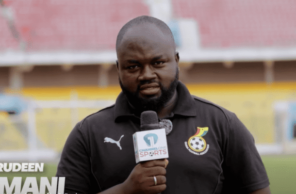 Male sports reporter holding a microphone in a stadium, wearing a black polo with a team crest; stadium seats in background and the channel logo on mic and shirt.