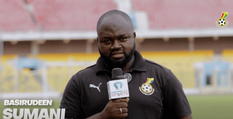 Male sports reporter holding a microphone in a stadium, wearing a black polo with a team crest; stadium seats in background and the channel logo on mic and shirt.