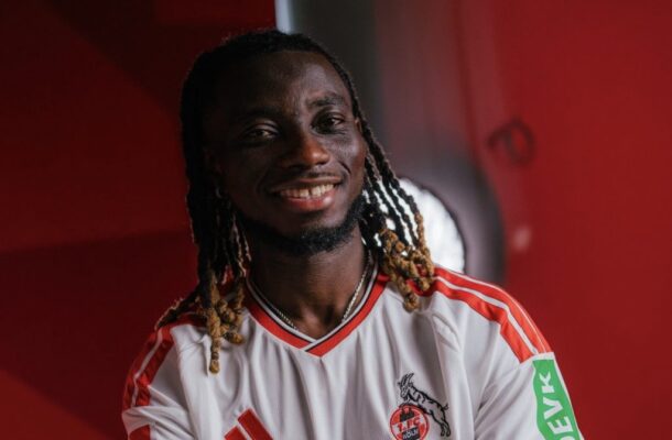 Smiling man with braided hair wearing a white jersey with red stripes, standing against a red background.