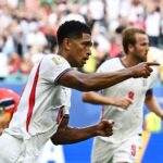 Two soccer players in white jerseys celebrate on the field as the cheering crowd watches in the background.