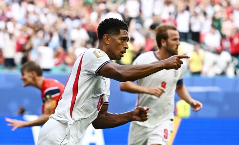 Two soccer players in white jerseys celebrate on the field as the cheering crowd watches in the background.