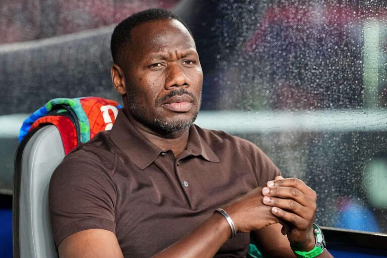 Man with a serious expression sits on a bus seat, hands clasped, rain streaks on the window behind him.