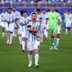 Argentina soccer player (captain) in white-and-sky striped kit crossing arms, center foreground on green field with teammates behind.