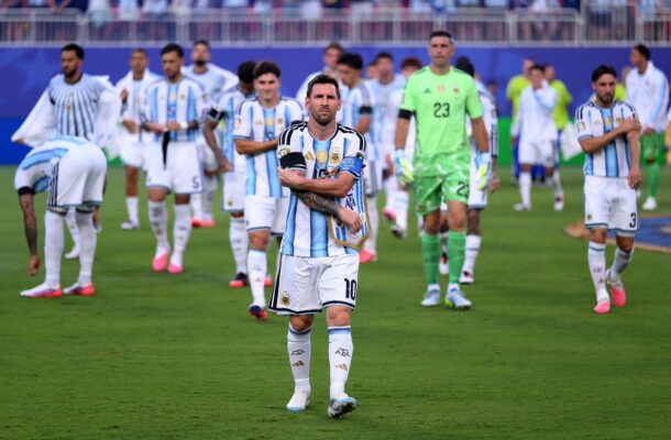 Argentina soccer player (captain) in white-and-sky striped kit crossing arms, center foreground on green field with teammates behind.