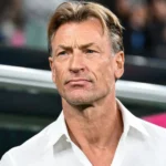 Close-up of a middle-aged man with blond hair in a white shirt, looking off to the side, with a blurred stadium background behind him.
