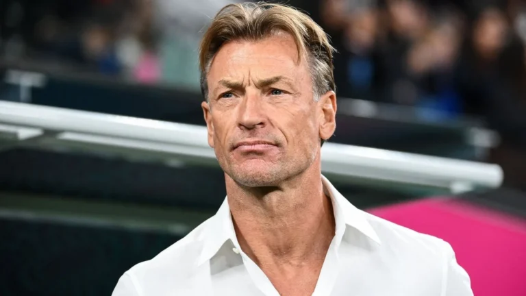 Close-up of a middle-aged man with blond hair in a white shirt, looking off to the side, with a blurred stadium background behind him.
