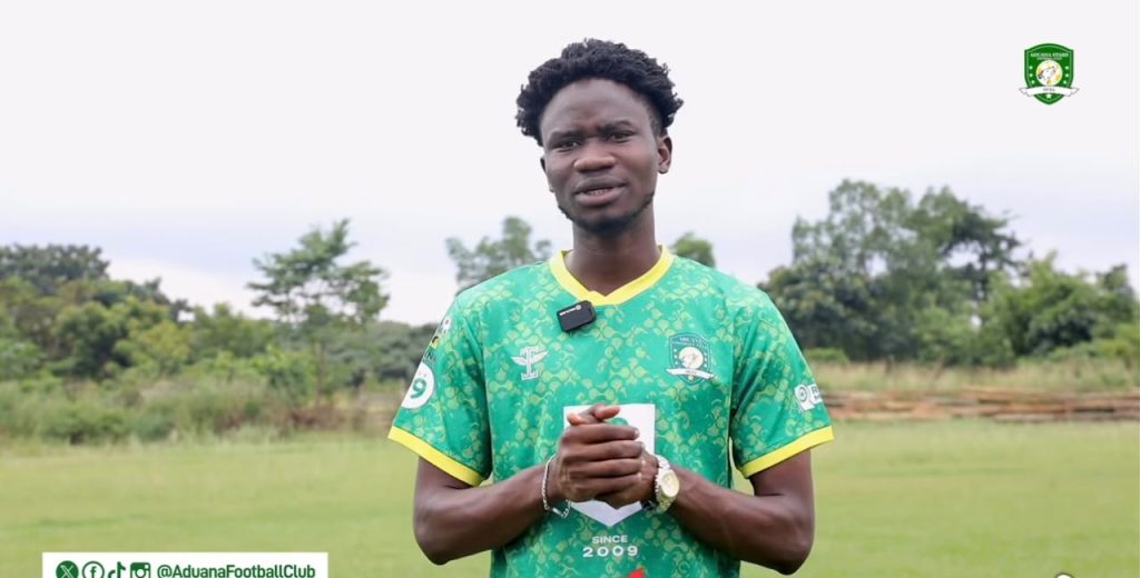 Young man in a green football jersey standing on a grassy field, hands clasped, with a club crest on his chest and a small microphone clipped to his shirt.