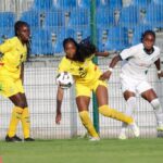 Three female soccer players in yellow and white uniforms contest a ball near a fence and blue stadium seats.