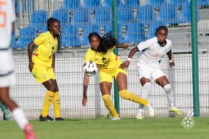 Three female soccer players in yellow and white uniforms contest a ball near a fence and blue stadium seats.