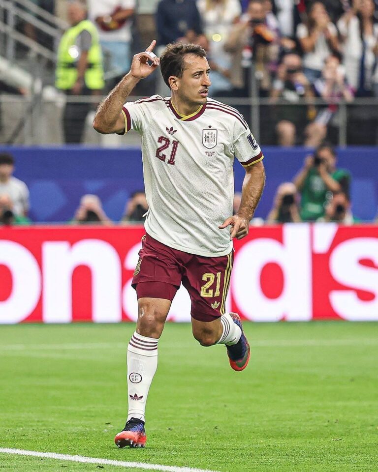 Soccer player in a white jersey with number 21 celebrating a goal on a green field, crowd in the stands behind him.