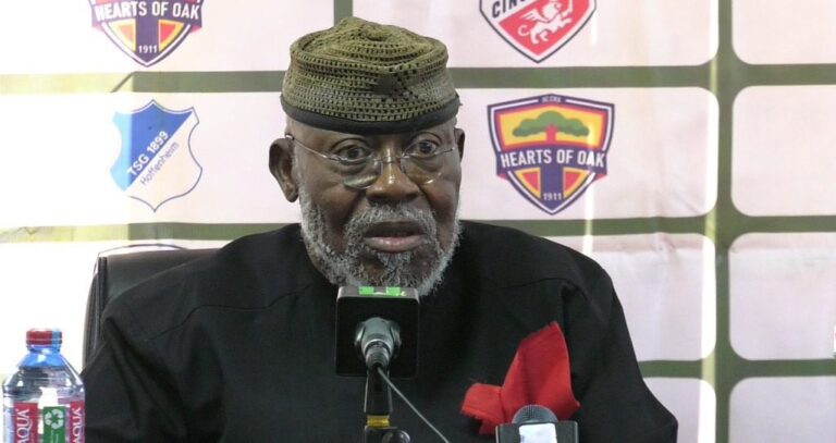 An older man wearing a traditional hat and glasses sits at a press conference, speaking into a microphone with Hearts of Oak logos in the backdrop.