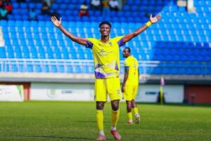 Soccer player in a yellow kit with arms raised in celebration on a green field at a stadium field side.