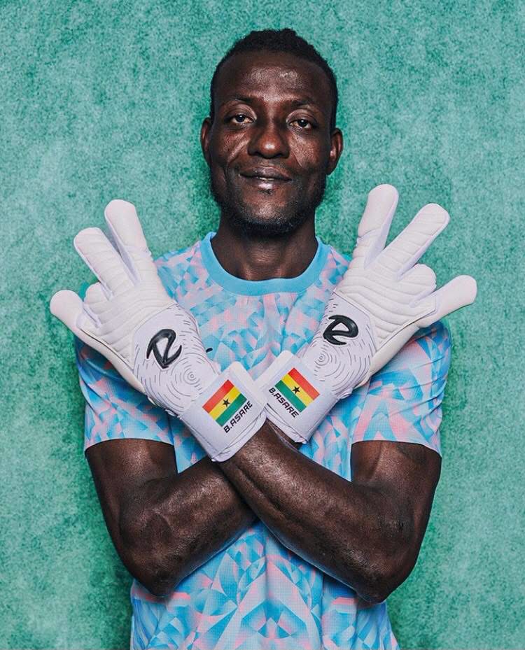 Goalkeeper posing with white gloves crossed over his chest, teal background, light blue patterned jersey; gloves show a Ghana flag and the name B. Asare on the wrist strap, conveying a sporting portrait.