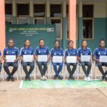 Group of twenty women in blue FIFA shirts sit in a row, each holding a certificate, during a coaching course ceremony outdoors in front of a green banner.