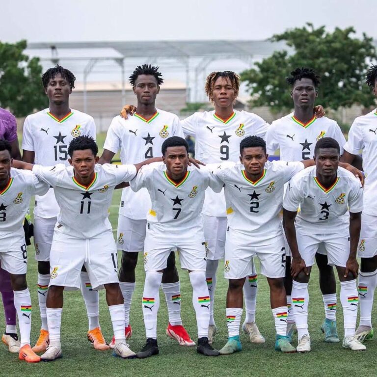 Group of soccer players in white uniforms posing for a team photo on a grass field, arms around shoulders, numbers visible on shirts.
