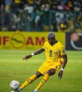 Soccer player in a yellow uniform (number 8) kicking a white ball on a green field, with spectators in the stands in the background.