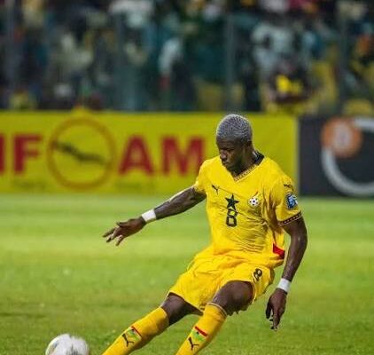 Soccer player in a yellow uniform (number 8) kicking a white ball on a green field, with spectators in the stands in the background.