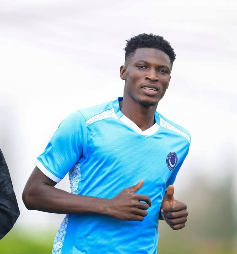 Young man in a light blue soccer jersey jogging outdoors, looking at the camera with a slight smile.