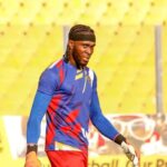Male football player in blue and red kit wearing gloves on a sunny stadium field, looking to the side during practice.
