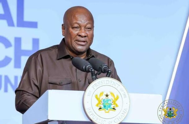 A man delivers a speech at a white podium with the Ghana presidency seal, two microphones, and a light blue backdrop behind him.