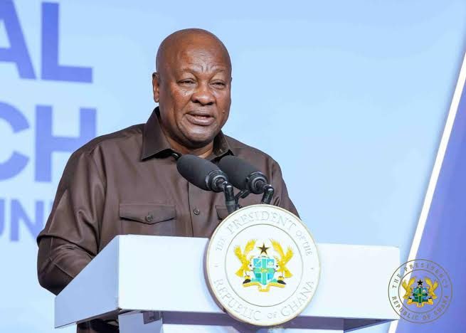 A man delivers a speech at a white podium with the Ghana presidency seal, two microphones, and a light blue backdrop behind him.