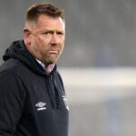 Man in a black Umbro hoodie on a sports field, looking serious and focused.