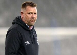 Man in a black Umbro hoodie on a sports field, looking serious and focused.