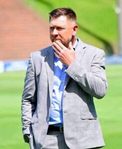 Man in a gray plaid suit stands outdoors, touching his chin in thought on a sunny day.