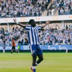 Soccer player in a blue-and-white striped kit with orange cleats raises arms in celebration on a green pitch as a full stadium cheers behind him.