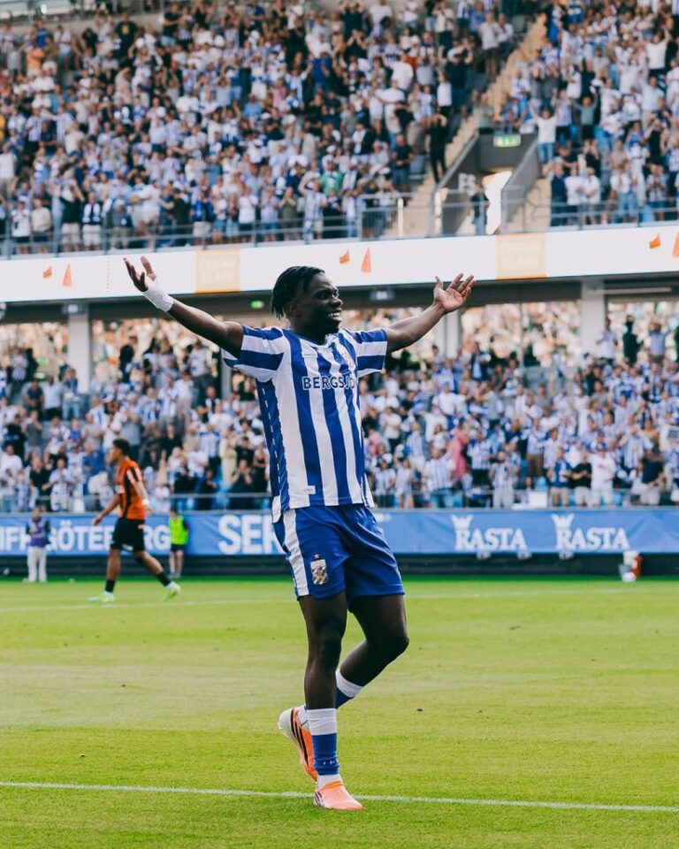 Soccer player in a blue-and-white striped kit with orange cleats raises arms in celebration on a green pitch as a full stadium cheers behind him.