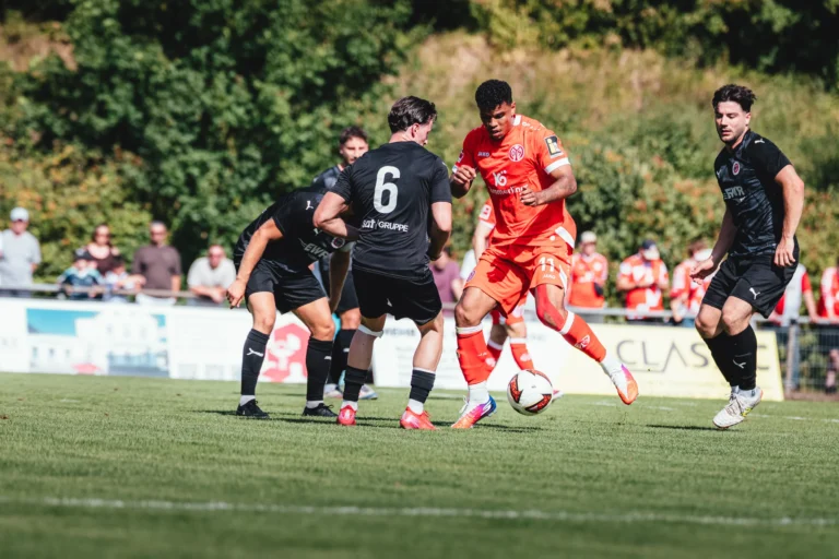 Soccer players in orange and black clash for the ball on a green field during a match, with spectators in the background.