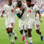 Three England players celebrate on the field, arms around each other in white jerseys with numbers 10 and 24, smiling after a goal.