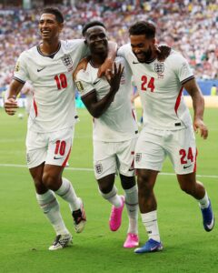 Three England players celebrate on the field, arms around each other in white jerseys with numbers 10 and 24, smiling after a goal.