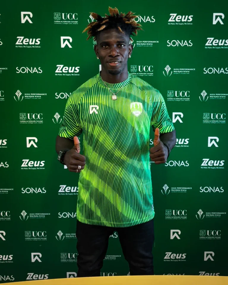 Athletic man in a neon green striped shirt giving two thumbs up, standing before a green sponsor backdrop with logos (Zeus, SONAS, UCC).