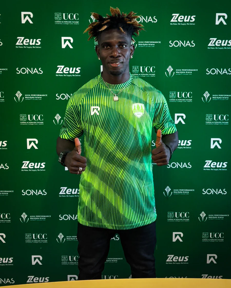 Athletic man in a neon green striped shirt giving two thumbs up, standing before a green sponsor backdrop with logos (Zeus, SONAS, UCC).