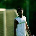 Athlete in a white sleeveless training shirt with green accents standing on a soccer field near a goal, looking off to the side.