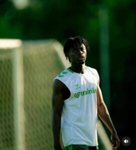 Athlete in a white sleeveless training shirt with green accents standing on a soccer field near a goal, looking off to the side.