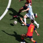 Soccer players in red and blue and light-blue uniforms chase a ball on a green field from an aerial perspective.