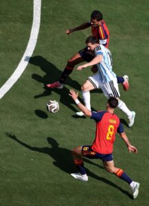 Soccer players in red and blue and light-blue uniforms chase a ball on a green field from an aerial perspective.