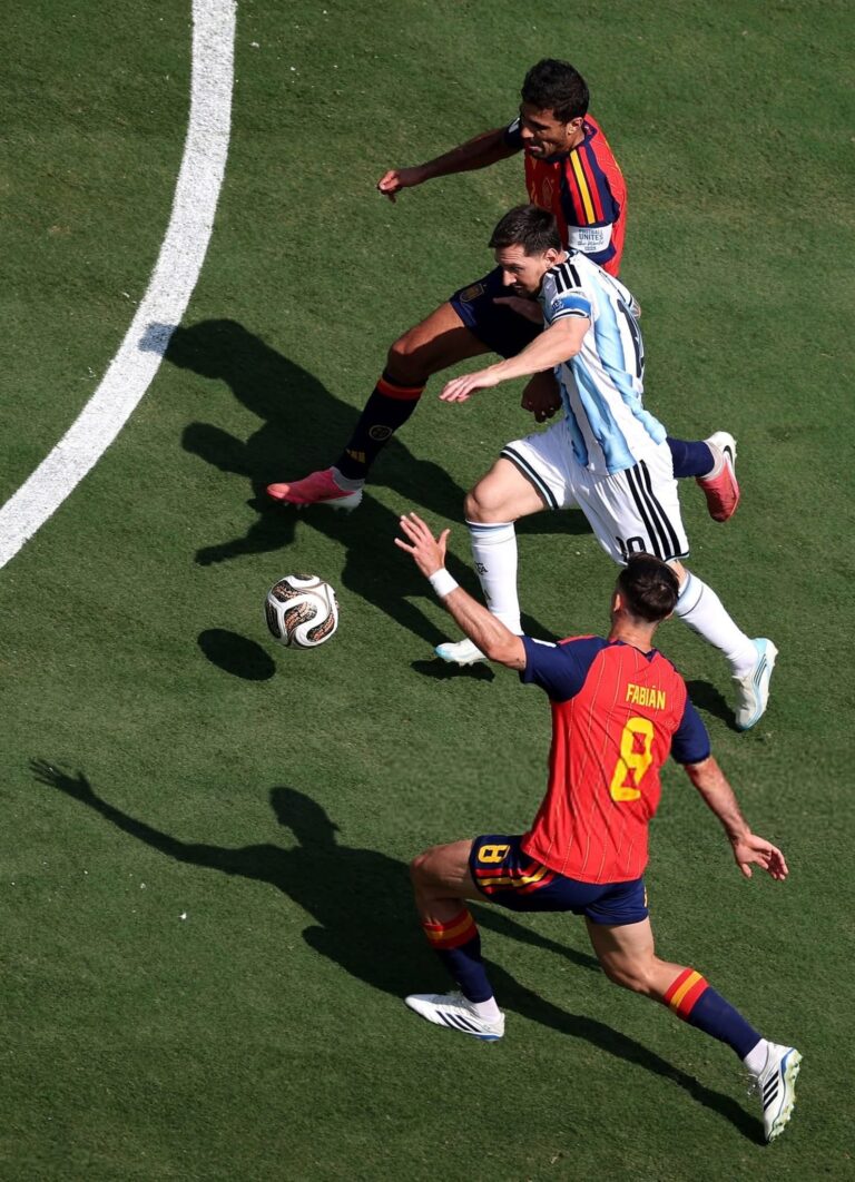 Soccer players in red and blue and light-blue uniforms chase a ball on a green field from an aerial perspective.