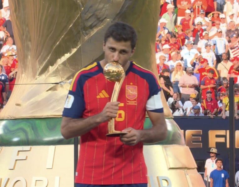 Spain national team player kissing the FIFA World Cup trophy on stage with a cheering crowd behind him.