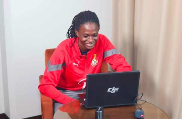 IMG_1336 | Ghanaguardian.com Person in a red athletic jacket with reflective stripes sits at a round table, smiling at a laptop.