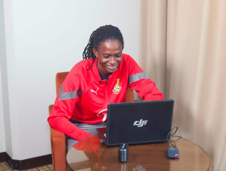 Person in a red athletic jacket with reflective stripes sits at a round table, smiling at a laptop.